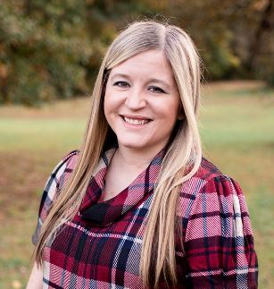 A white person with long, blonde hair smiling at the camera. She is wearing a red and white plaid shirt.
