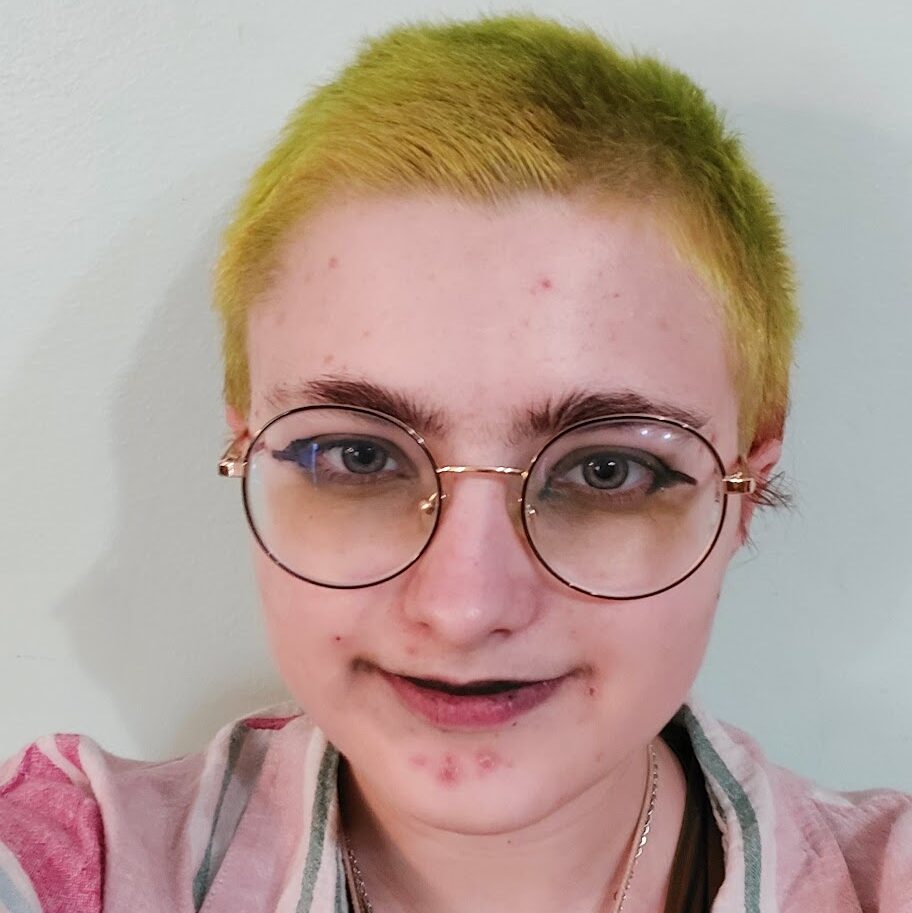 A white person with round glasses and short, yellow hair taking a selfie. Xe wears a pink and blue striped button up over a black tanktop.