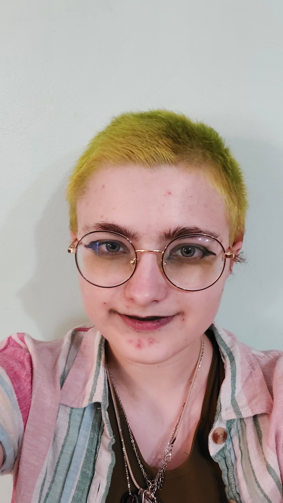A white person with round glasses and short, yellow hair taking a selfie. Xe wears a pink and blue striped button up over a black tanktop.