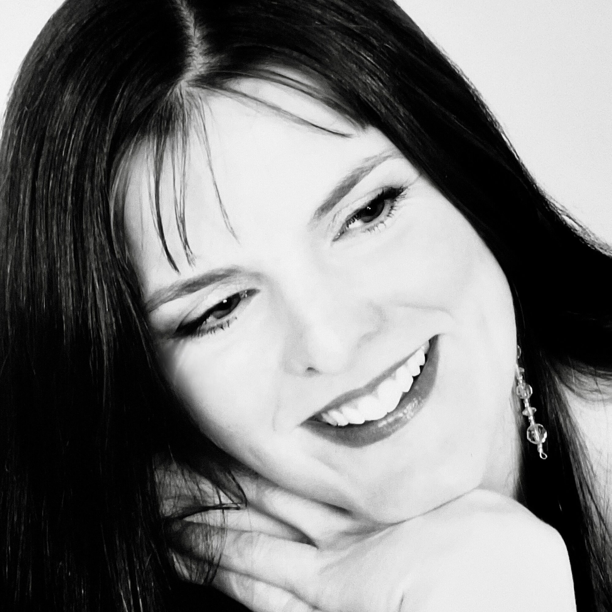 The photo is in black and white and depicts a white person with long, dark hair smiling to the right of the shot.
