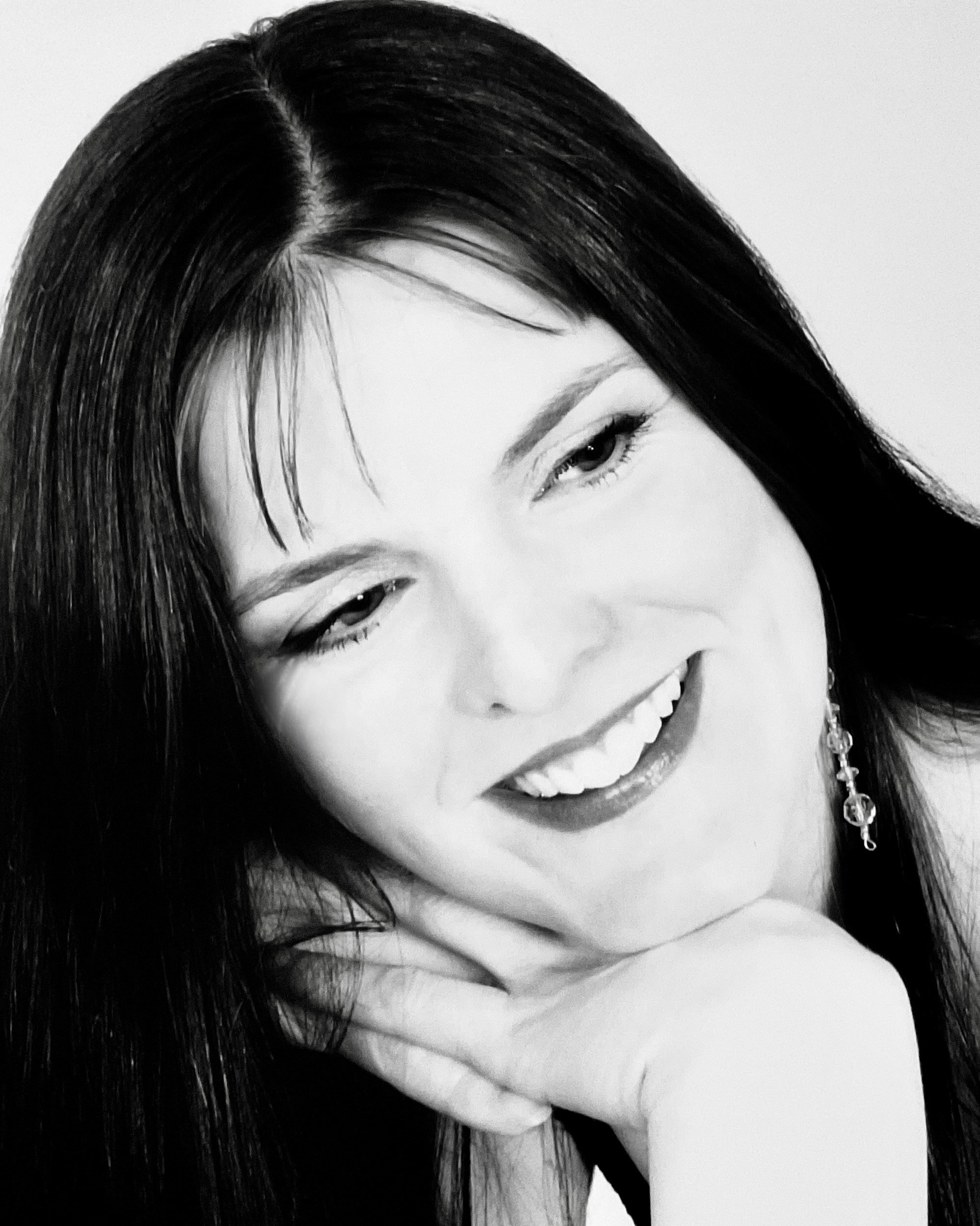 The photo is in black and white and depicts a white person with long, dark hair smiling to the right of the shot.