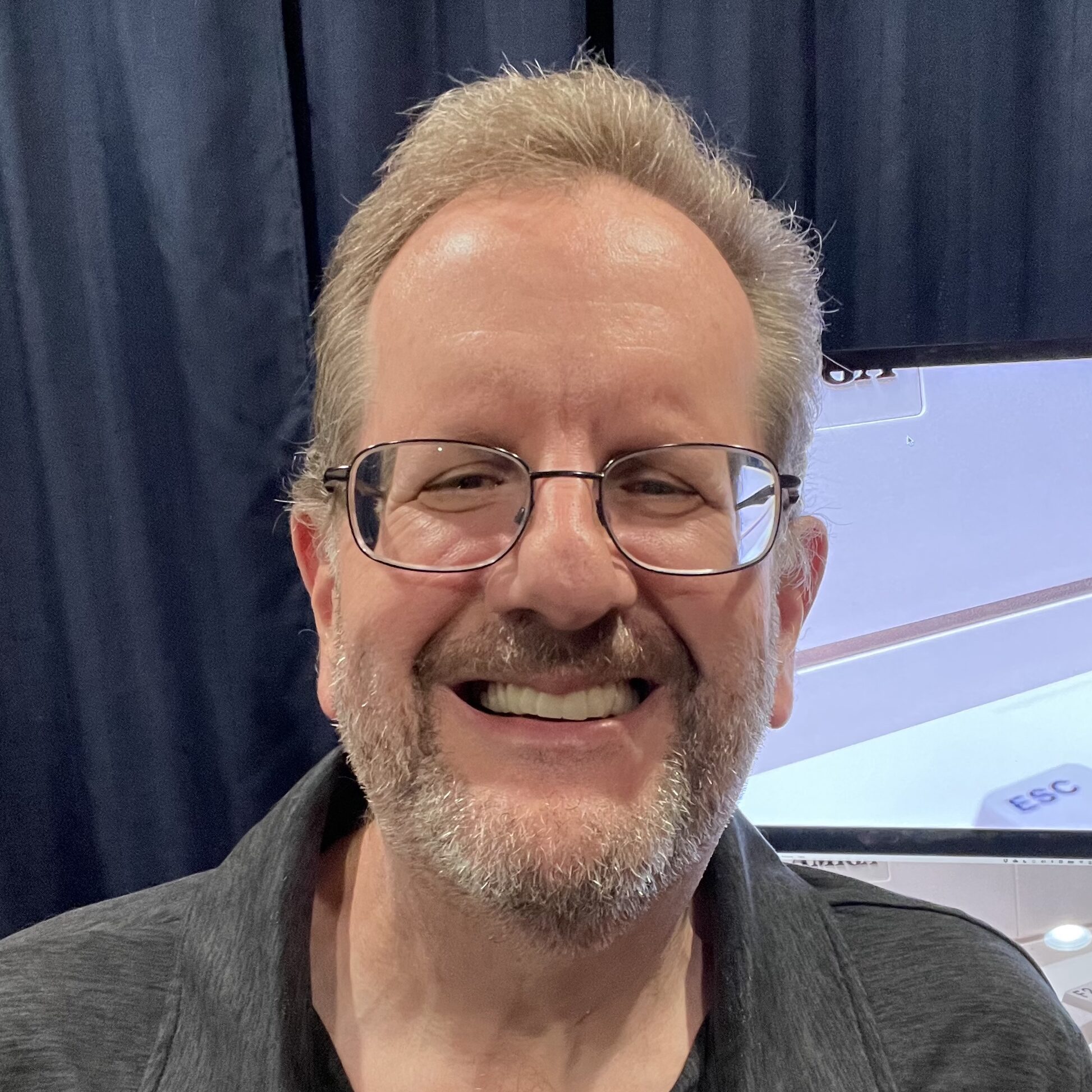 A white man with gray hair and a beard smiles at the camera. He wears wire-rimmed glasses and a dark gray shirt.