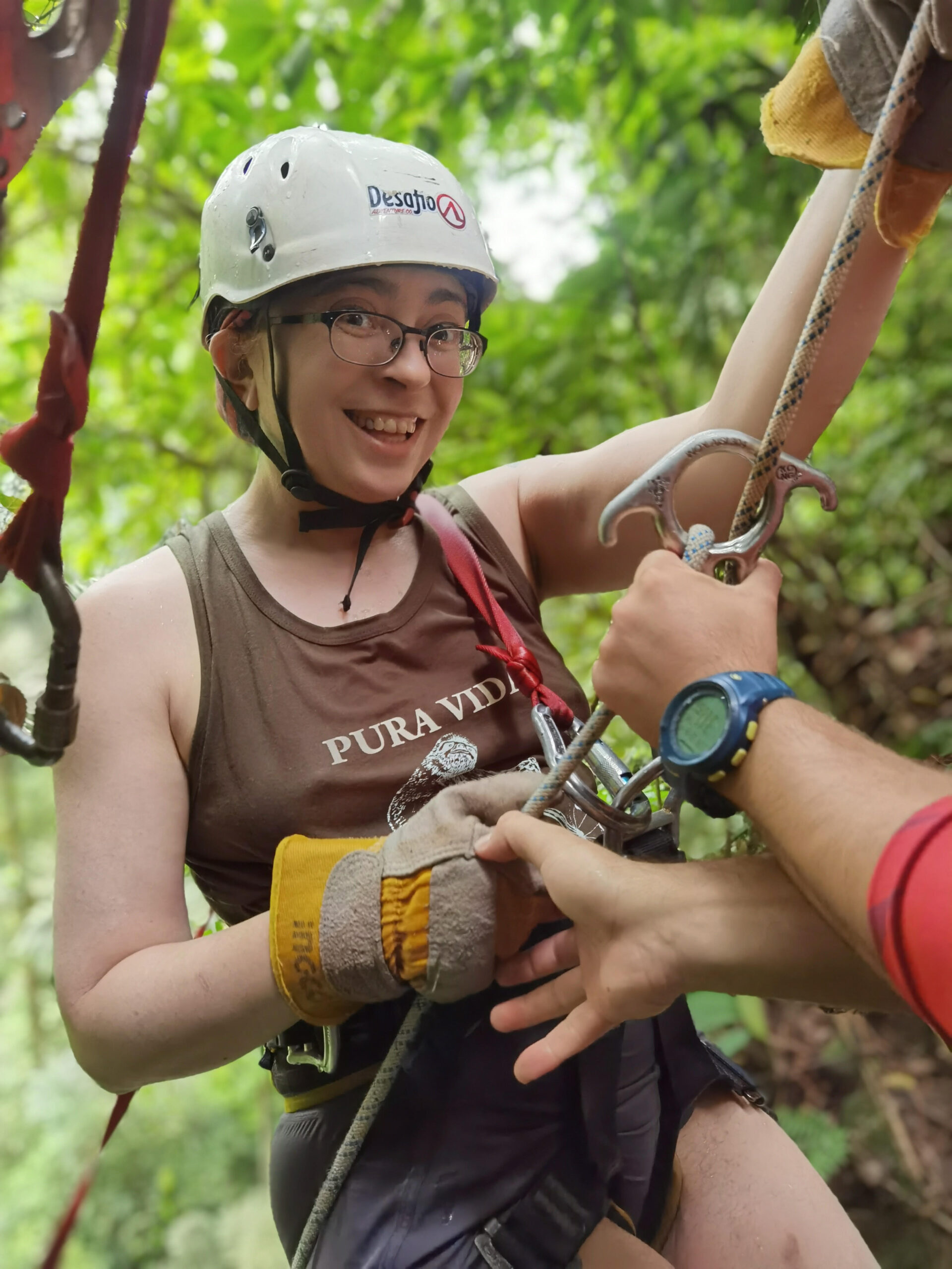 A person in a brown tanktop and a white helmet climbing a rope and smiling at the camera.