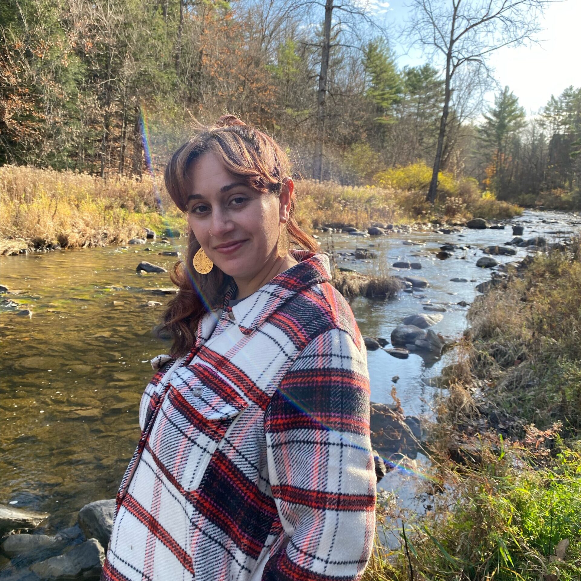 A woman with brown skin and long, brown hair stands in front of a creek, looking at the camera. She wears a plaid red and white shirt.