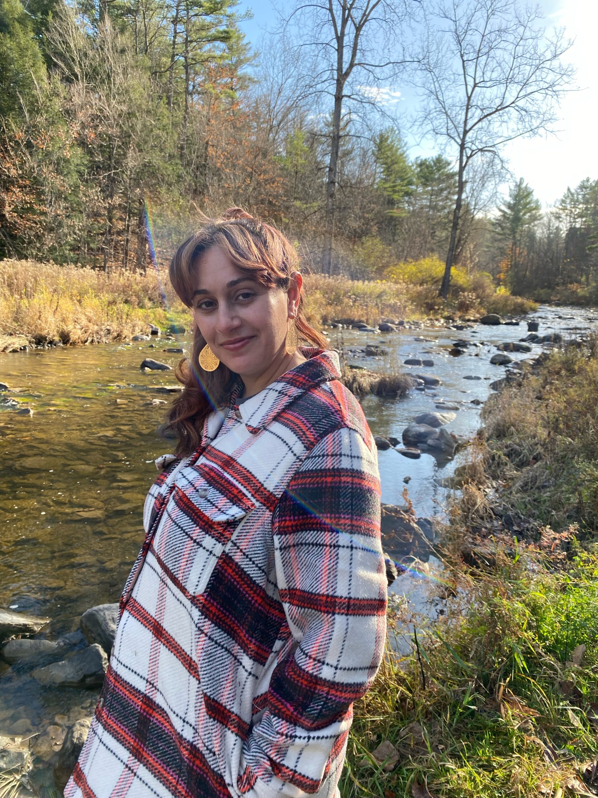 A woman with brown skin and long, brown hair stands in front of a creek, looking at the camera. She wears a plaid red and white shirt.