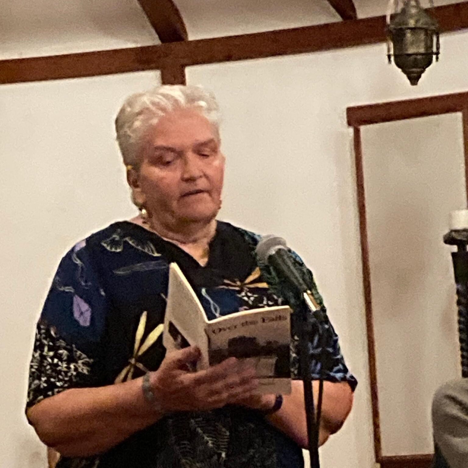 A white woman with short, white hair reads a book at a microphone.