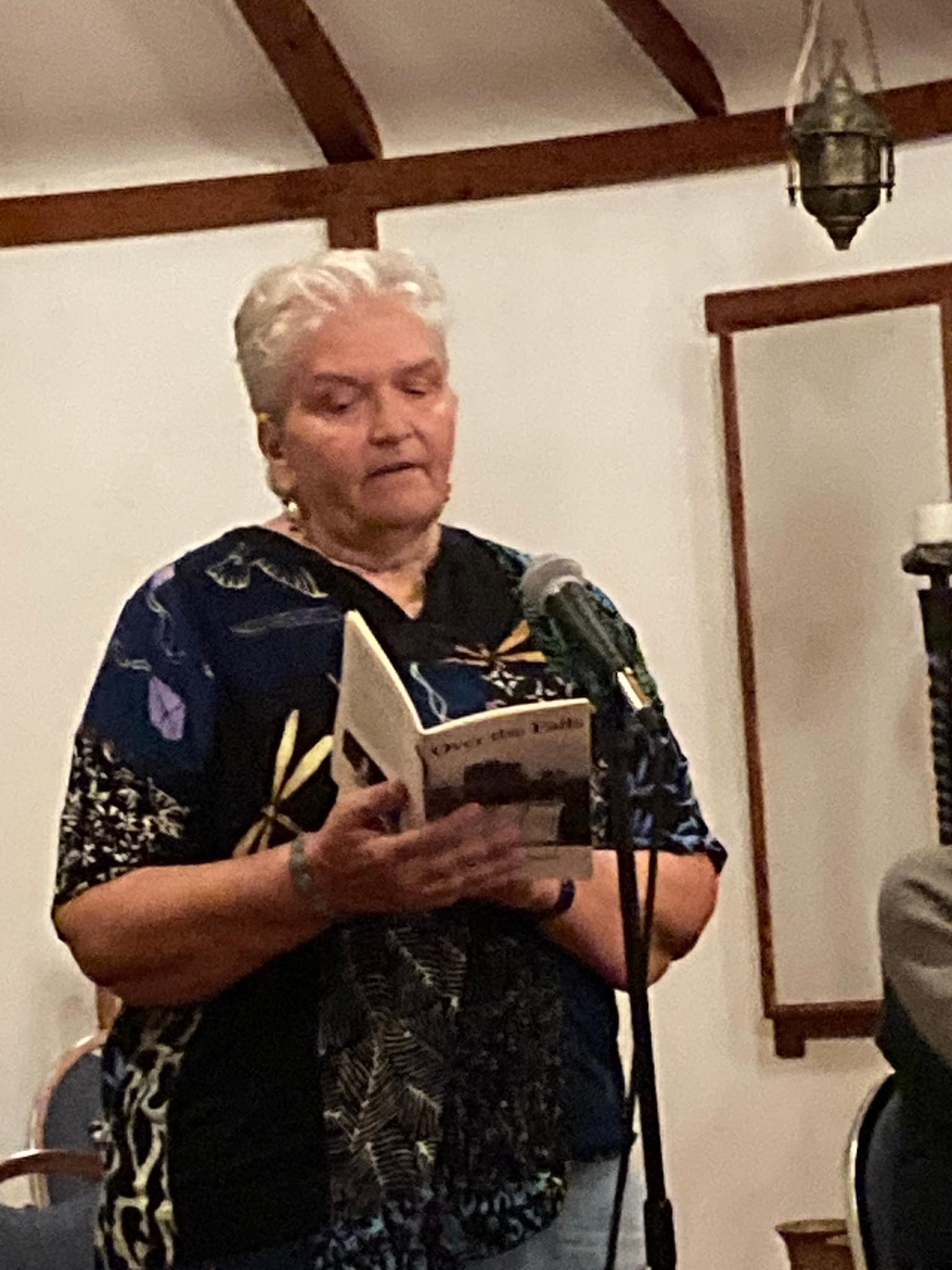 A white woman with short, white hair reads a book at a microphone.