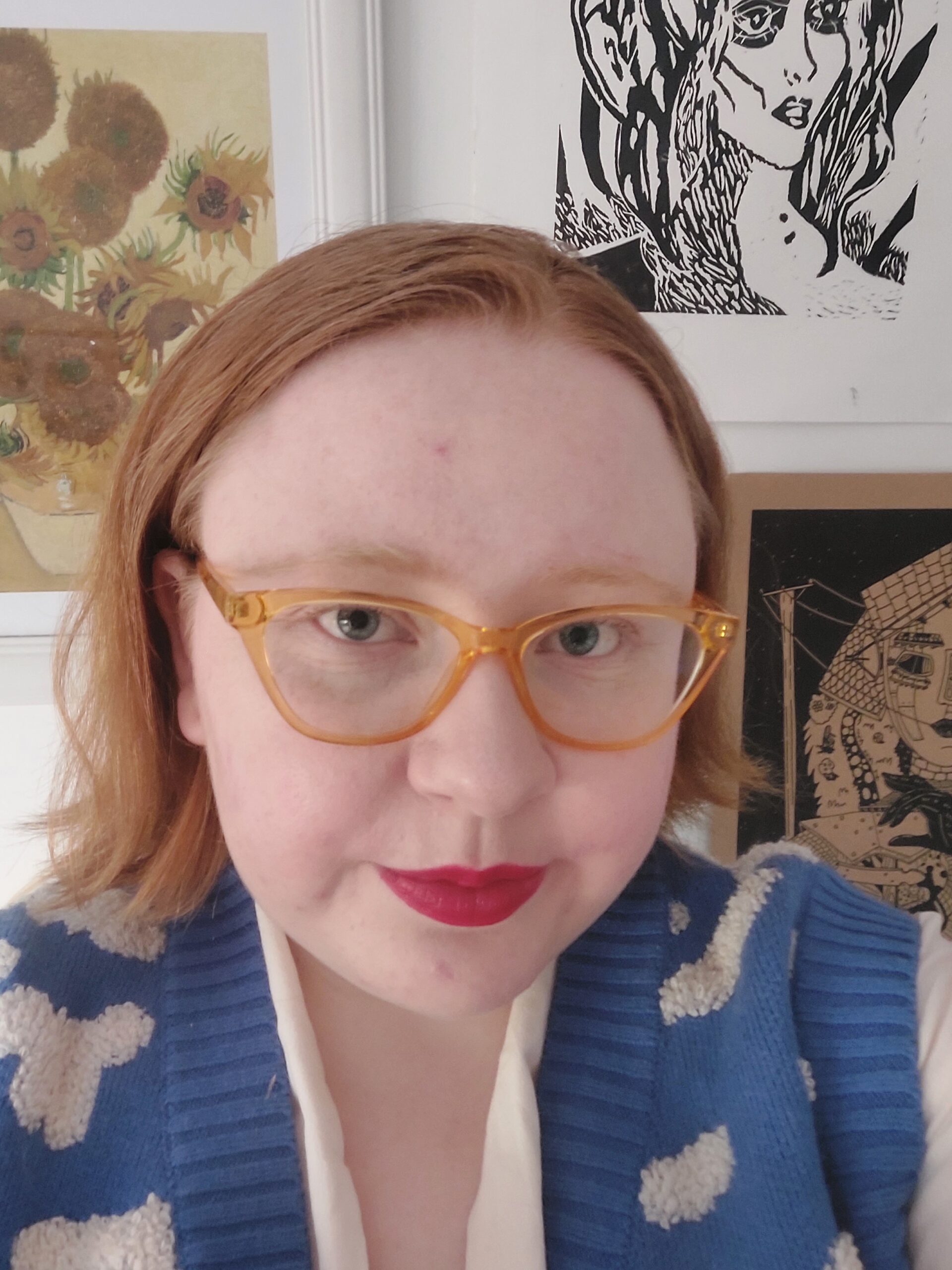 A pale, blonde person wearing red lipstick, orange-rimmed glasses, and a blue sweater with white clouds on it. She is standing against the backdrop of several pieces of art.