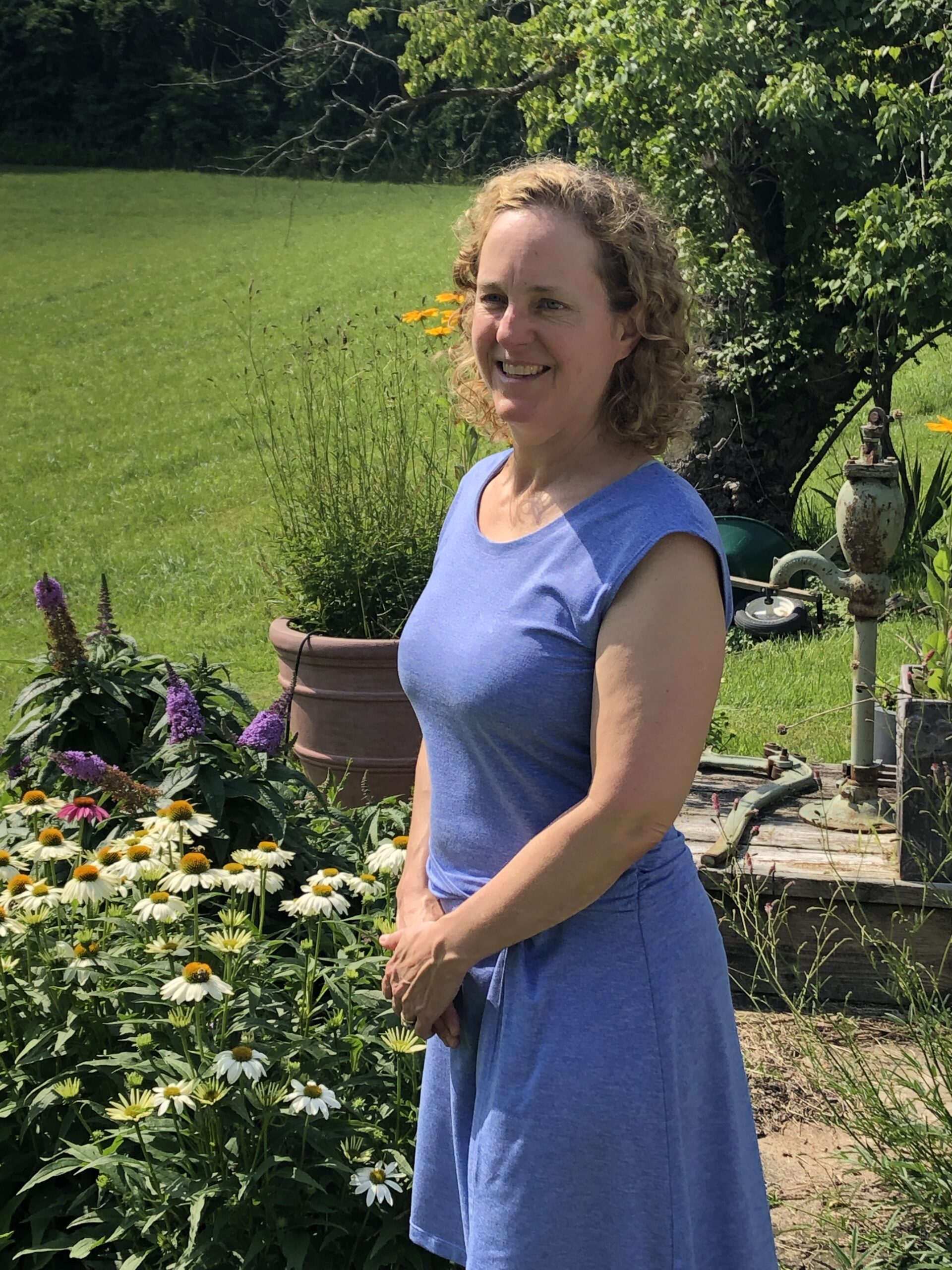 A white woman with shoulder-length light brown hair stands in a garden. She is smiling and wearing a blue dress with short sleeves.