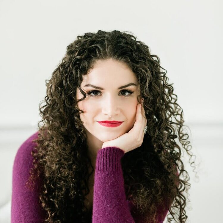 A white woman with long, curly, brown hair. She is wearing a purple sweater and we can see her from the waist up. The background is white.