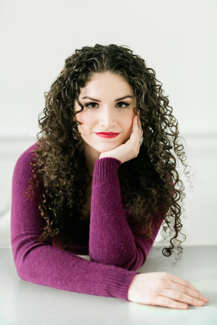 A white woman with long, curly, brown hair. She is wearing a purple sweater and we can see her from the waist up. The background is white.