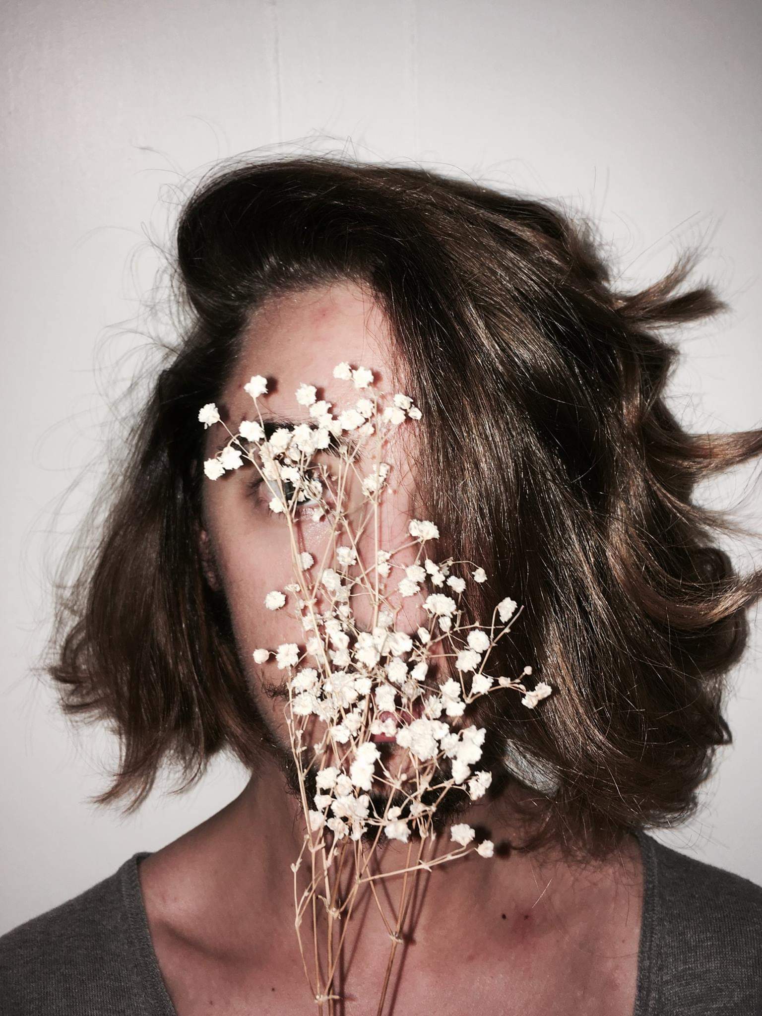 A person with shoulder-length black hair. He is hiding behind a small bunch of white flowers.