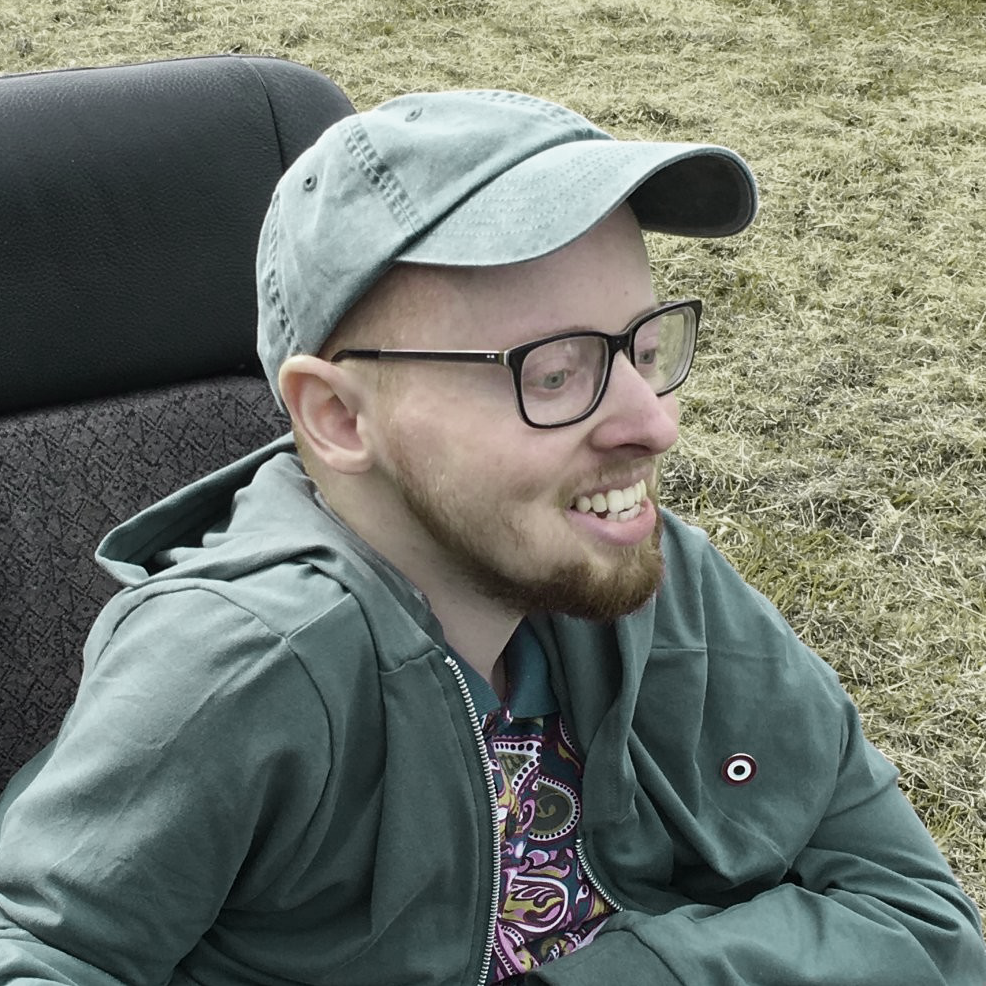A white man sits in a black wheelchair and smiles to something off-camera. He wears a blue baseball hat and a green sweatshirt.