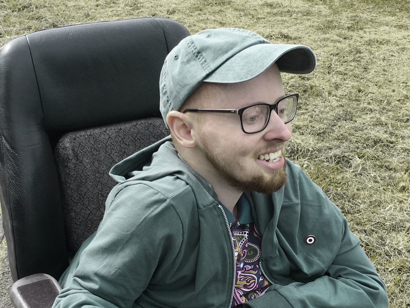 A white man sits in a black wheelchair and smiles to something off-camera. He wears a blue baseball hat and a green sweatshirt.