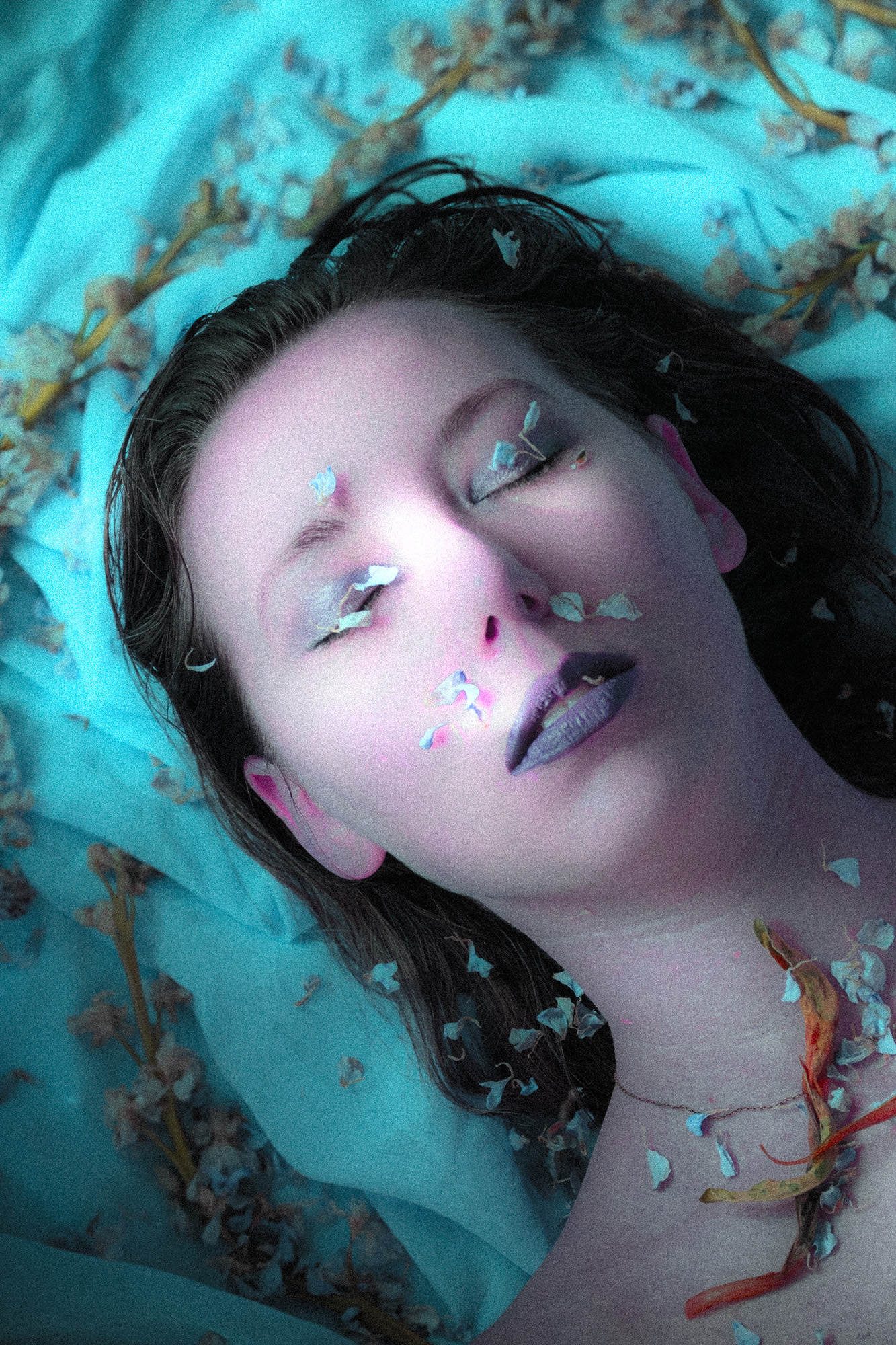 A white woman lays on the ground with her eyes closed. She is covered in turquoise petals. The fabric she lays on is also turquoise and covered in flowers.
