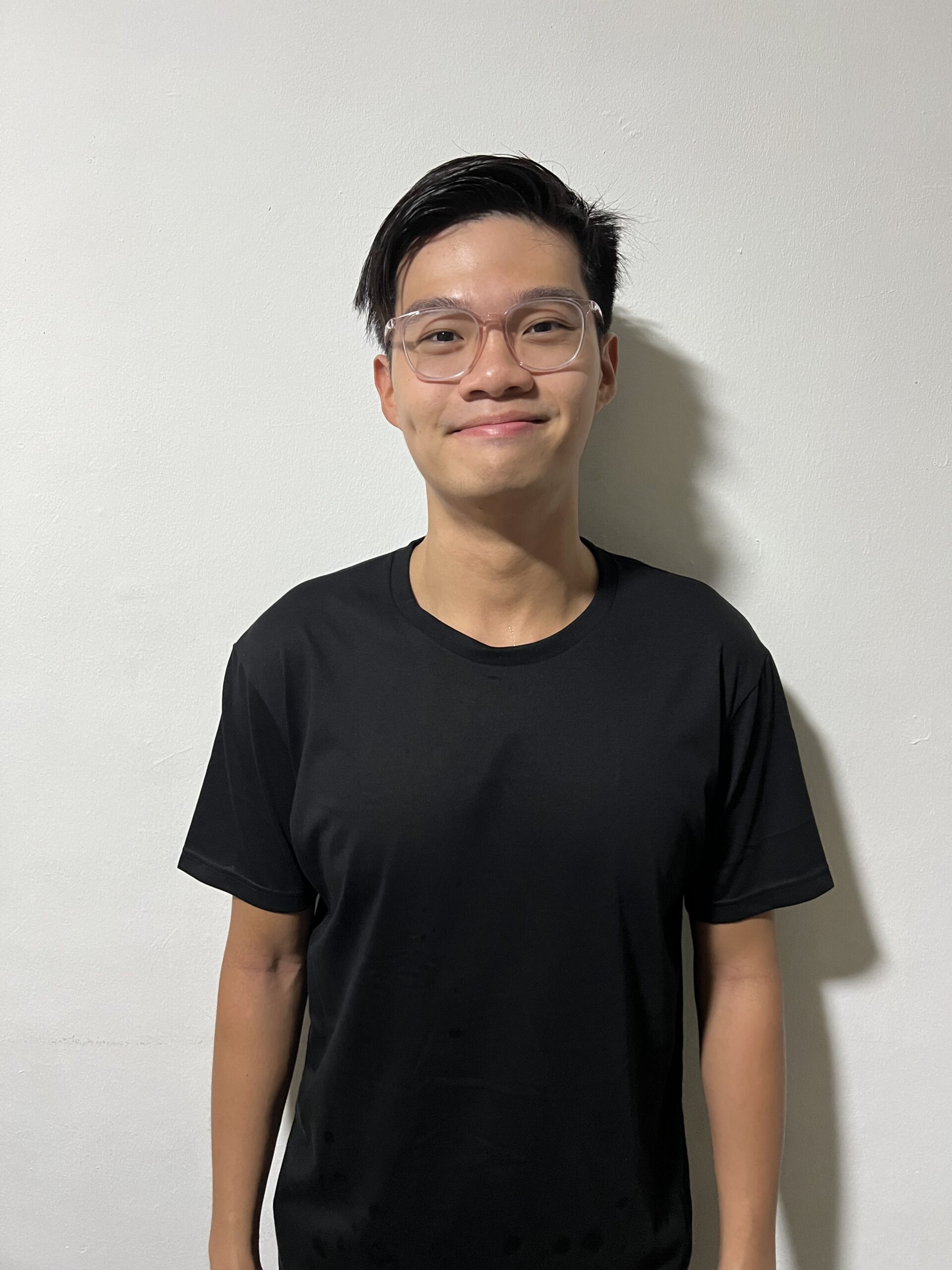 An Asian man smiling with his mouth closed and looking at the camera. He has short, black hair and is wearing a black shirt and glasses. He is standing against a white wall.