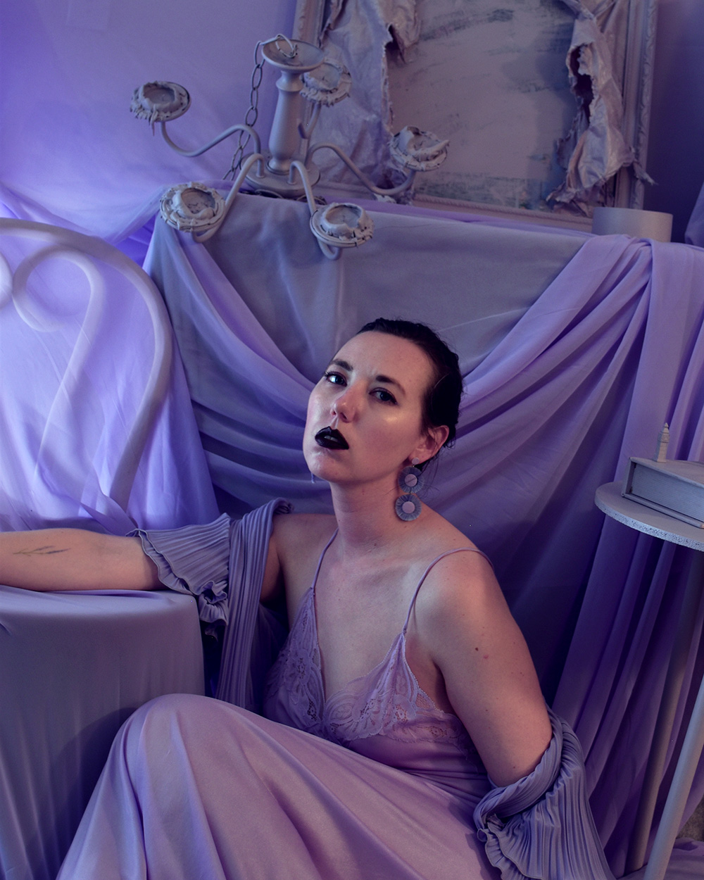 A white woman poses for the camera, crouching. She wears a lilac dress and earings and dark purple lipstick and is surrounded by lilac furnature.