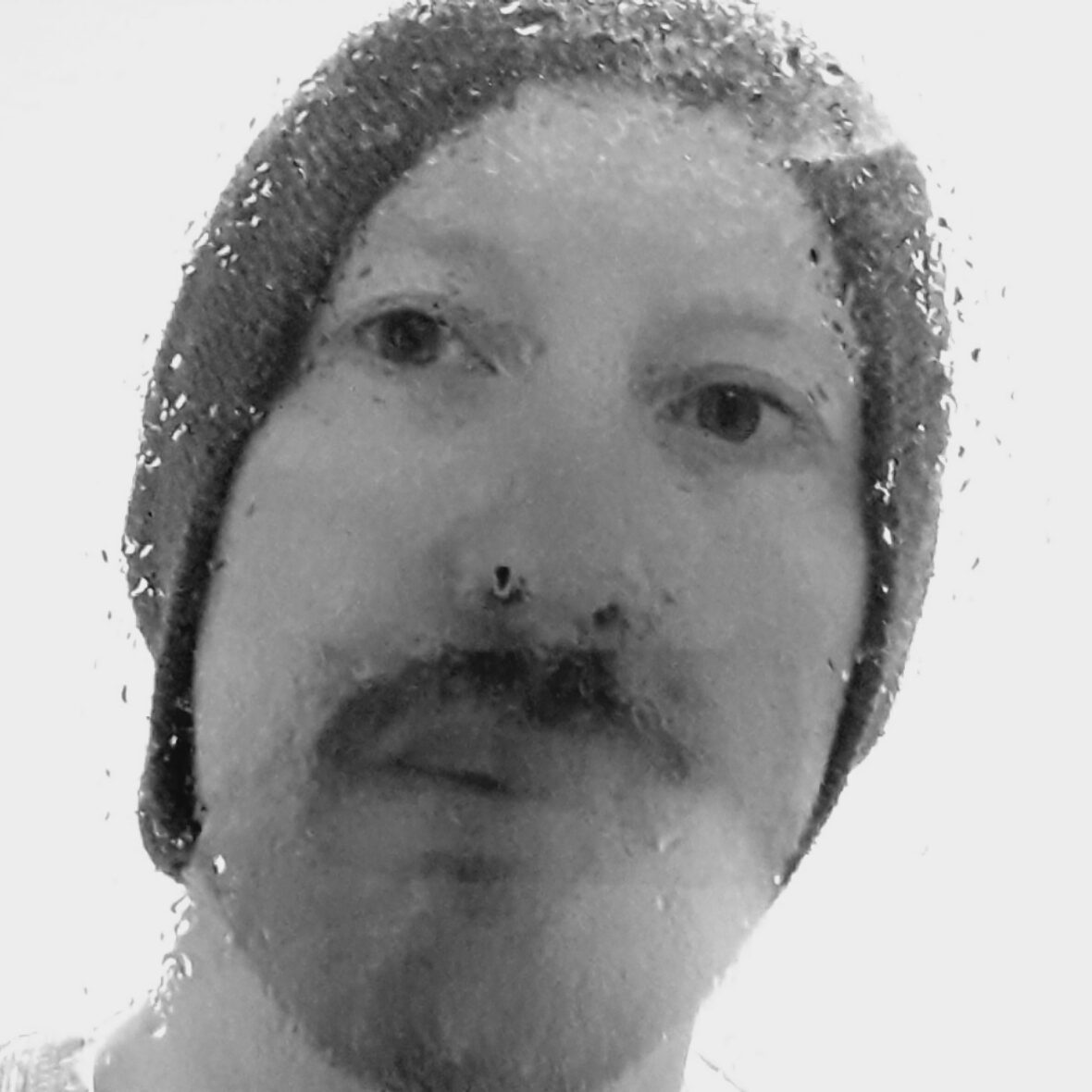 A photo in black and white. A white man wearing a hat with a moustache gazes at the camera. There are water droplets on the lens.