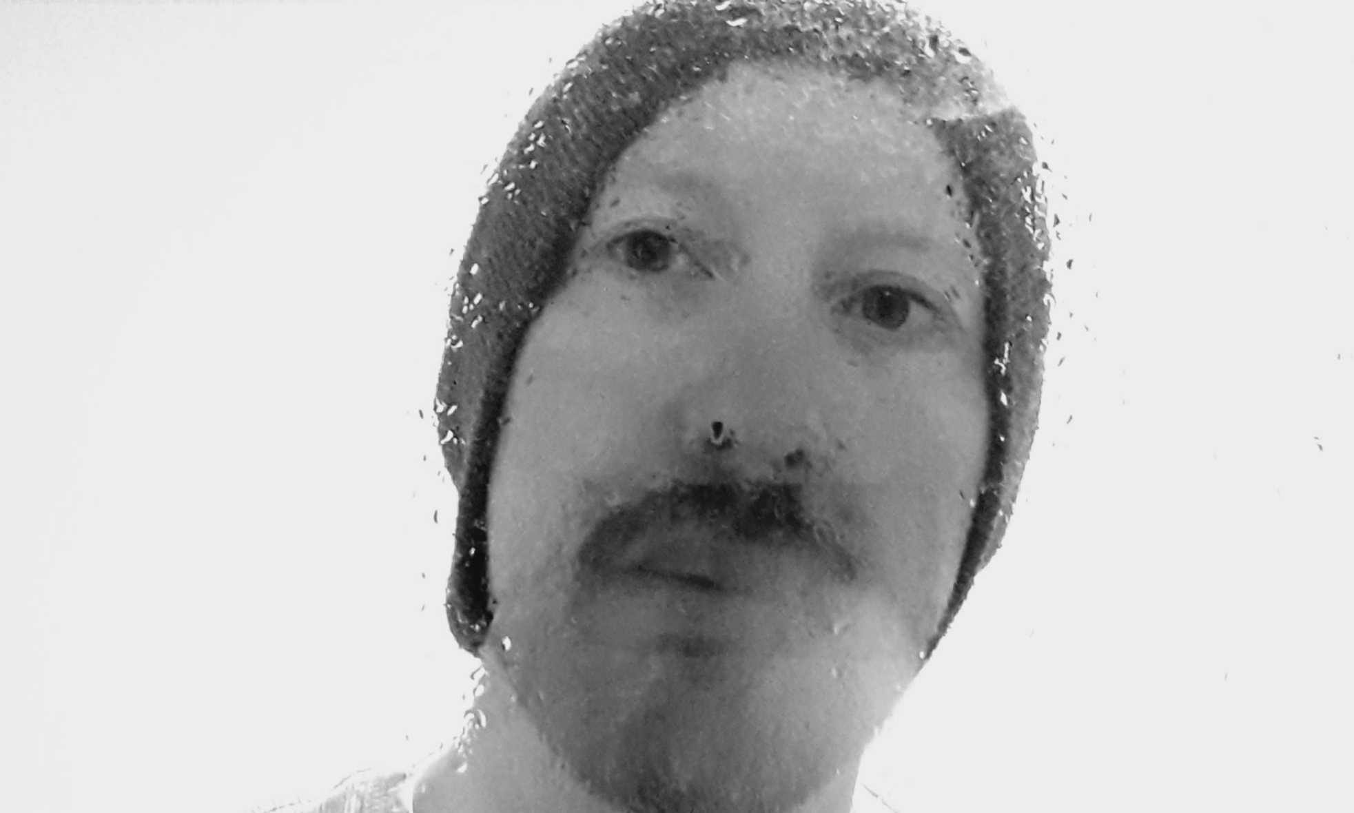 A photo in black and white. A white man wearing a hat with a moustache gazes at the camera. There are water droplets on the lens.