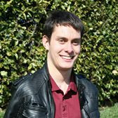 A white man with dark hair wearing a black jacket over a red button up. He is standing in front of a leafy green backdrop.