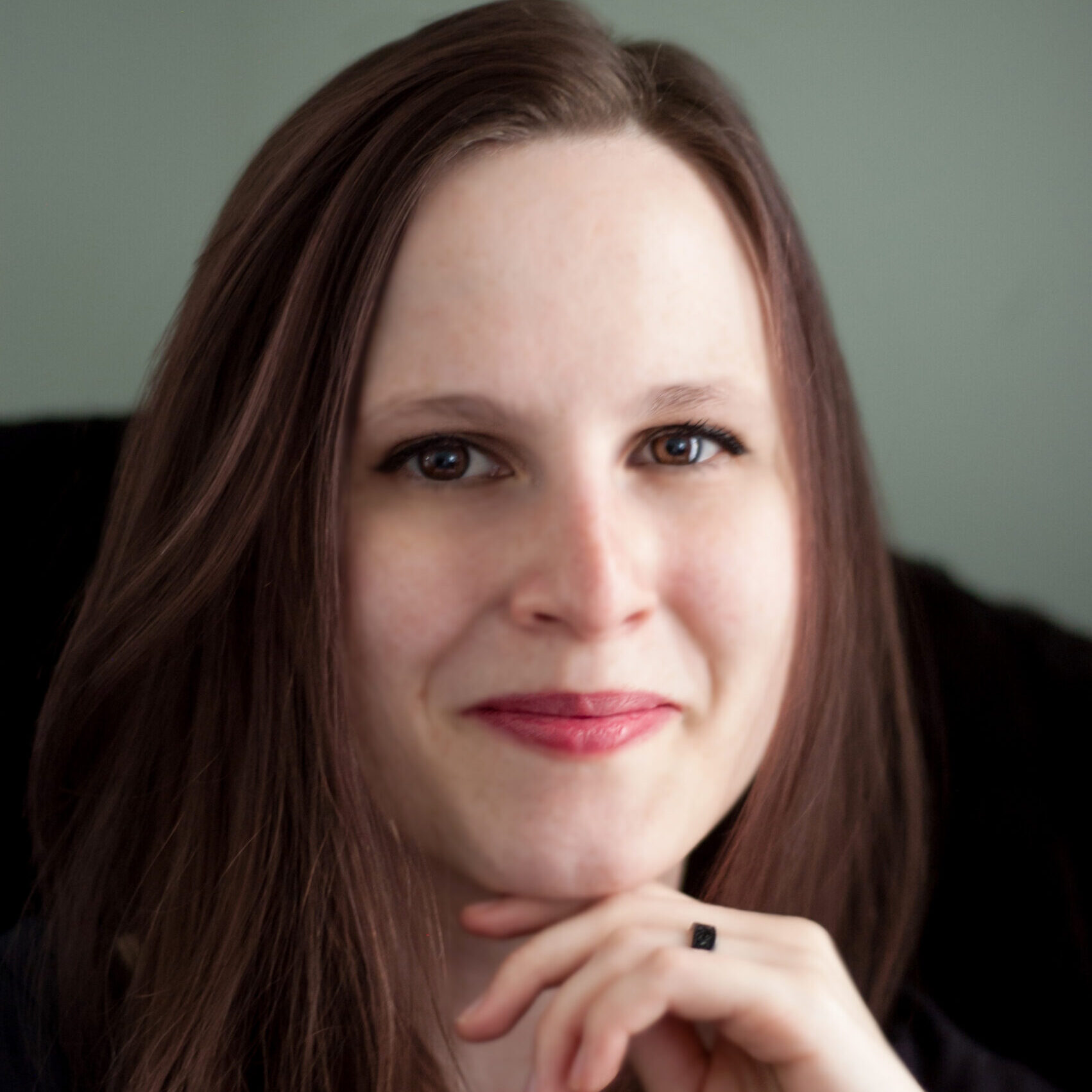 A white woman smiling with her mouth closed. She has long, chestnut hair and wears pink lipstick.