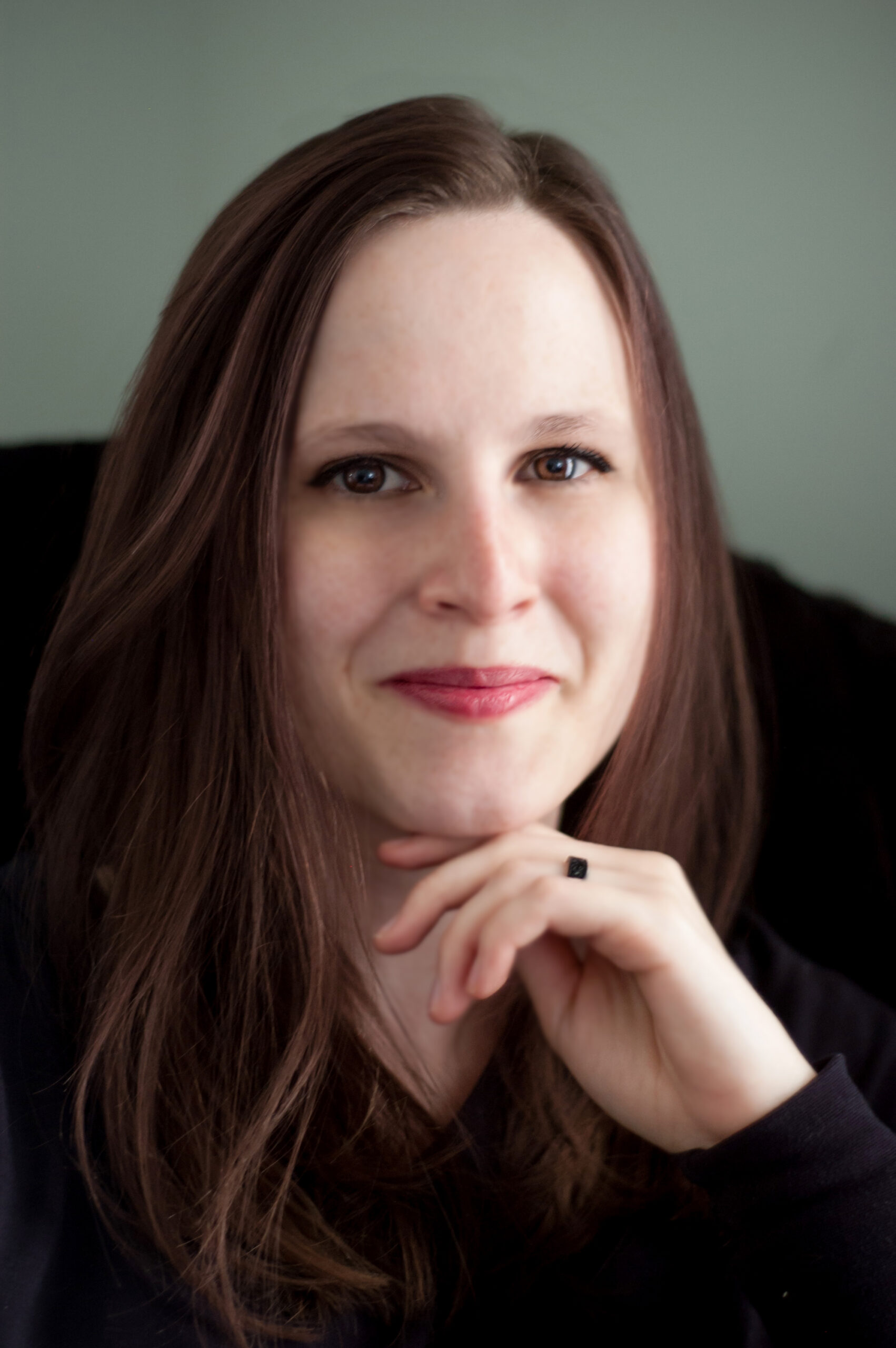 A white woman smiling with her mouth closed. She has long, chestnut hair and wears pink lipstick.