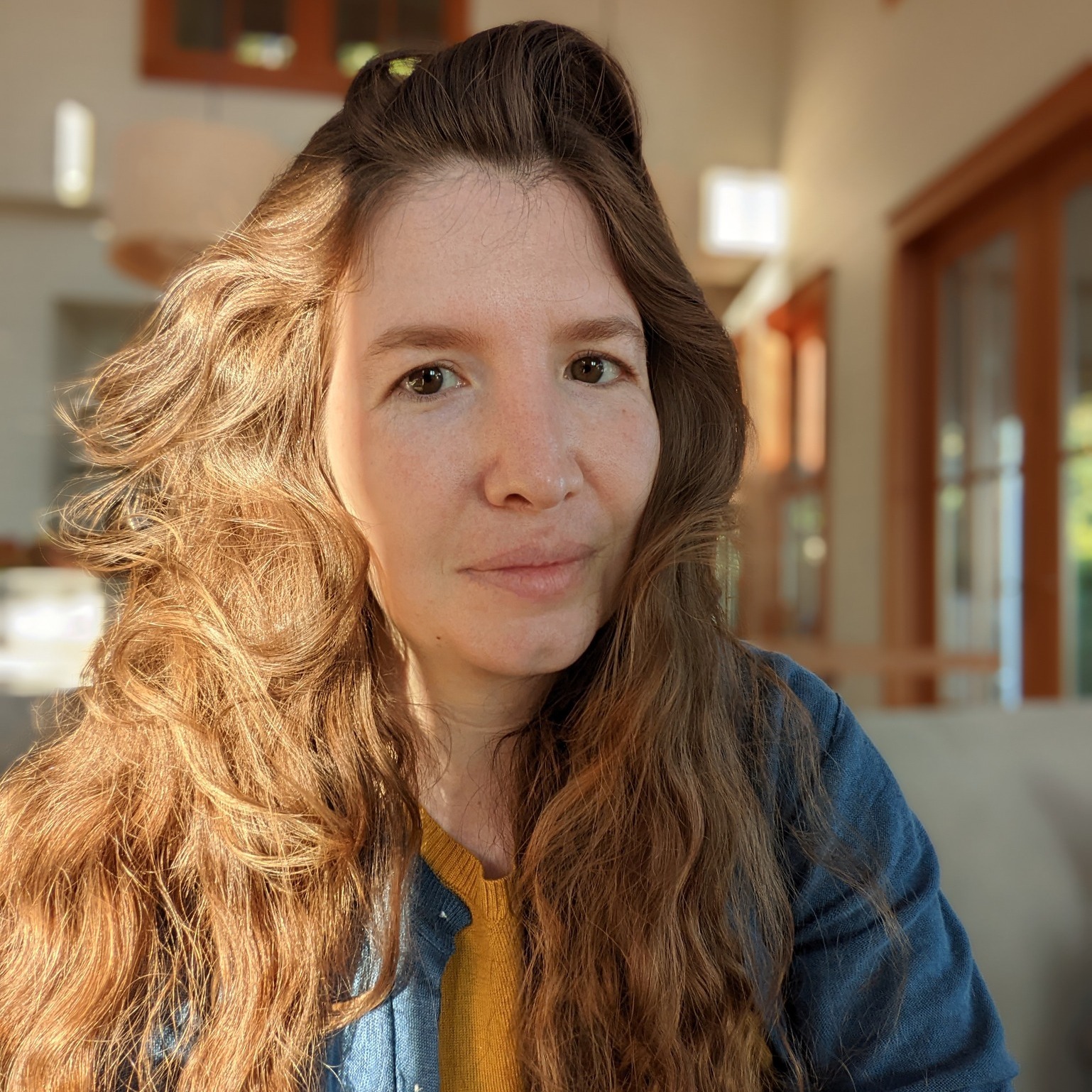 A white woman with long, brown hair gazes at the camera. She is indoors and the background is blurred. She wears a blue jacket over a yellow sweater.