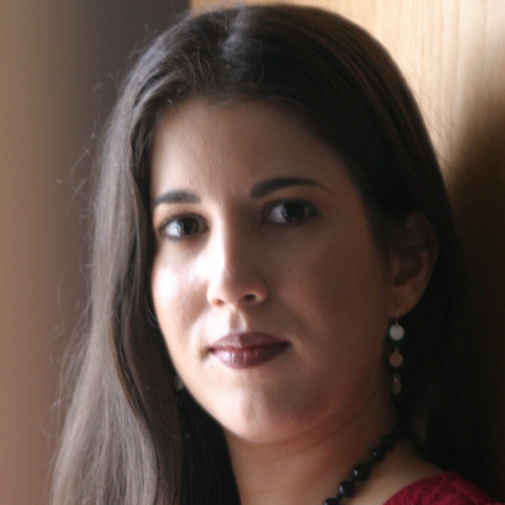 A woman with brown skin and long, brown hair gazes at the camera. She wears a pink shirt and a beaded black necklace.