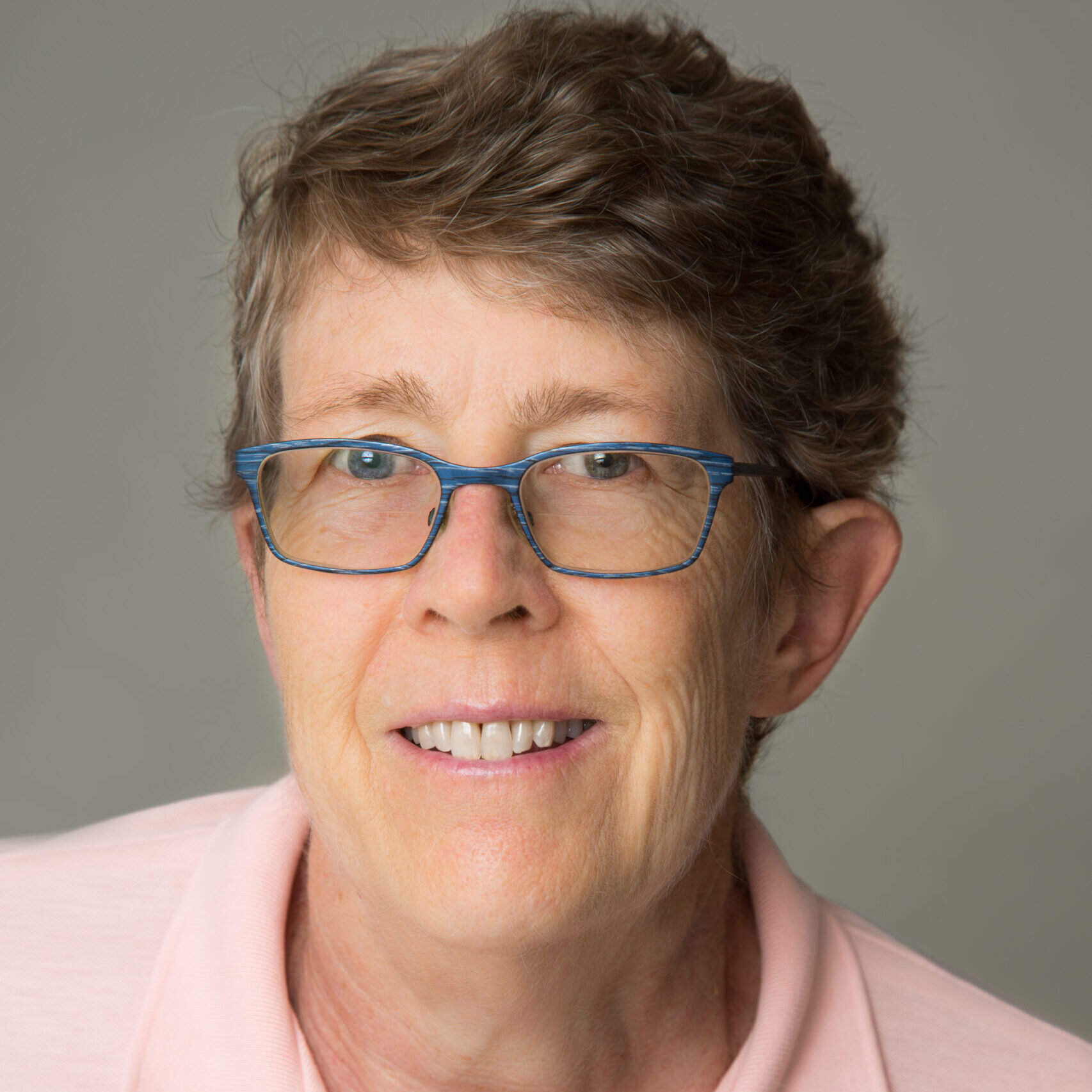 A white woman with short, light brown hair smiles at the camera. She wears blue-rimmed glasses and a pink polo shirt.