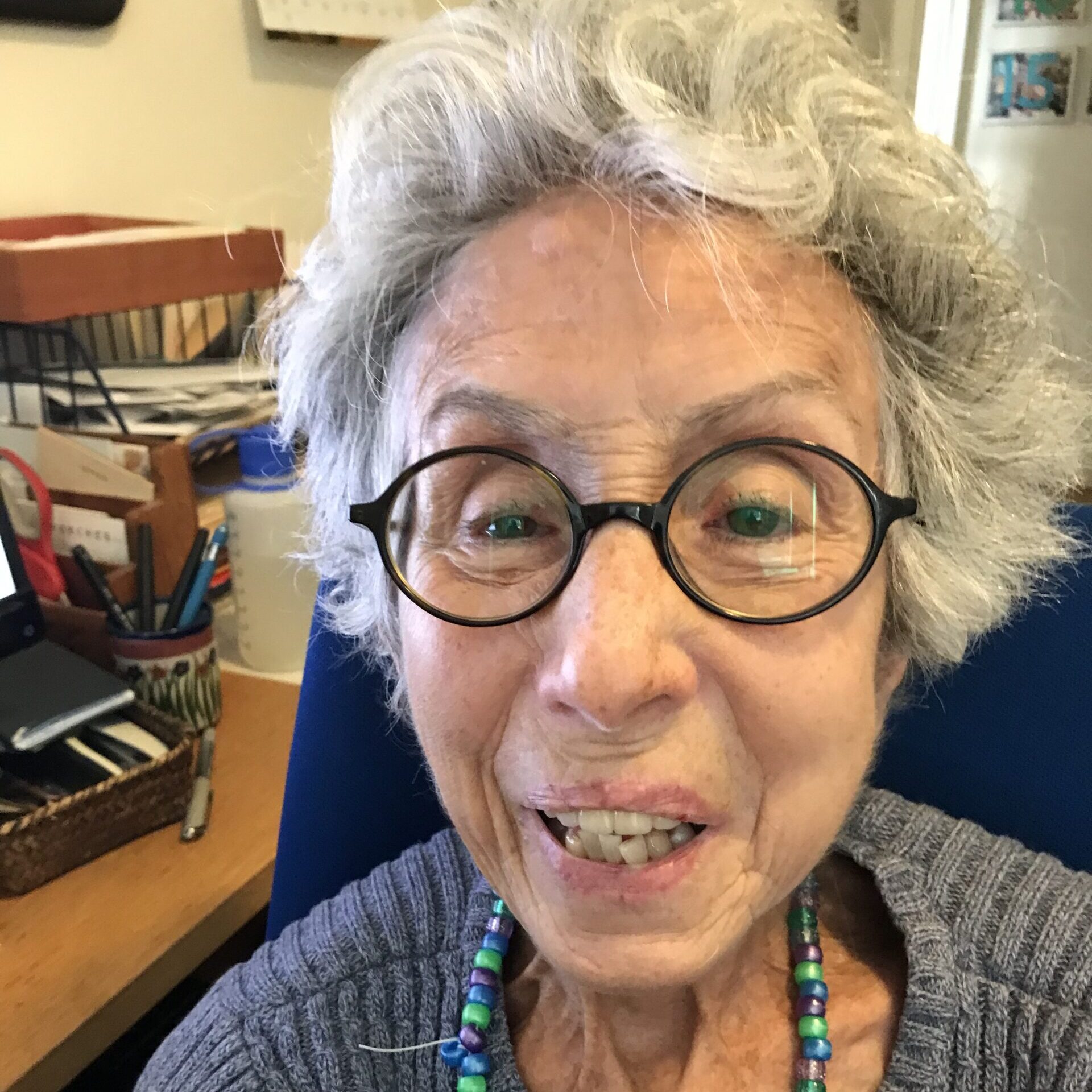 A woman with short, gray hair smiles at the camera. She wears round, black-rimmed glasses; a green, beaded necklace; and a blue-gray sweater.