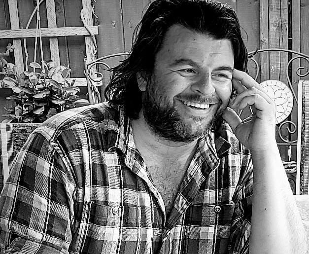 A black and white photo of a white man in a button-up flannel smiling at the camera. He is sitting outside on a bench. His hair is dark and shoulder-length and he has a short beard.