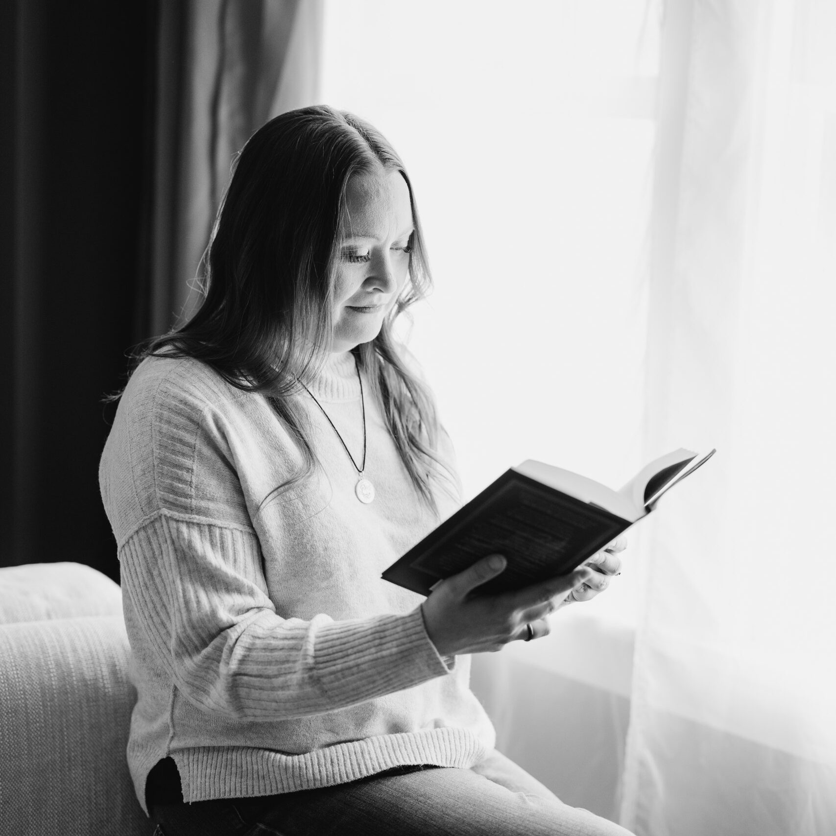 A black and white photo of a woman with long hear reading a book. She is sitting by a window and wears a light-colored sweater.