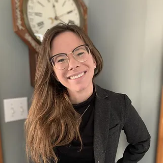 A white woman with long, light brown hair wearing a black jacket and green pants. She smiles at the camera and stands in front of a large clock.