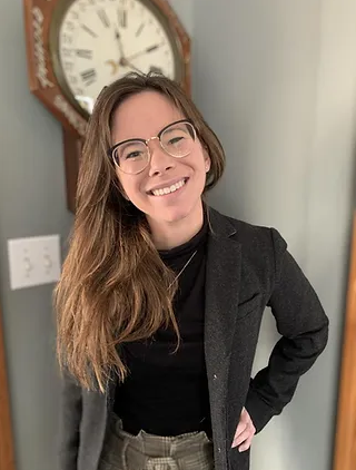 A white woman with long, light brown hair wearing a black jacket and green pants. She smiles at the camera and stands in front of a large clock.