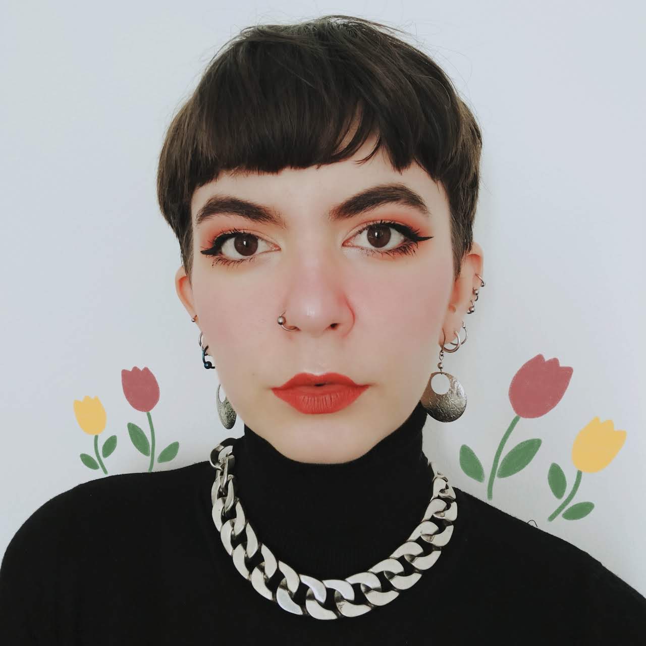 A white woman with short, brown hair gazes at the camera. She wears bright red lipstick, dangly silver earrings, and a black turtleneck sweater. The background is a white wall with yellow and pink tulips painted on it.