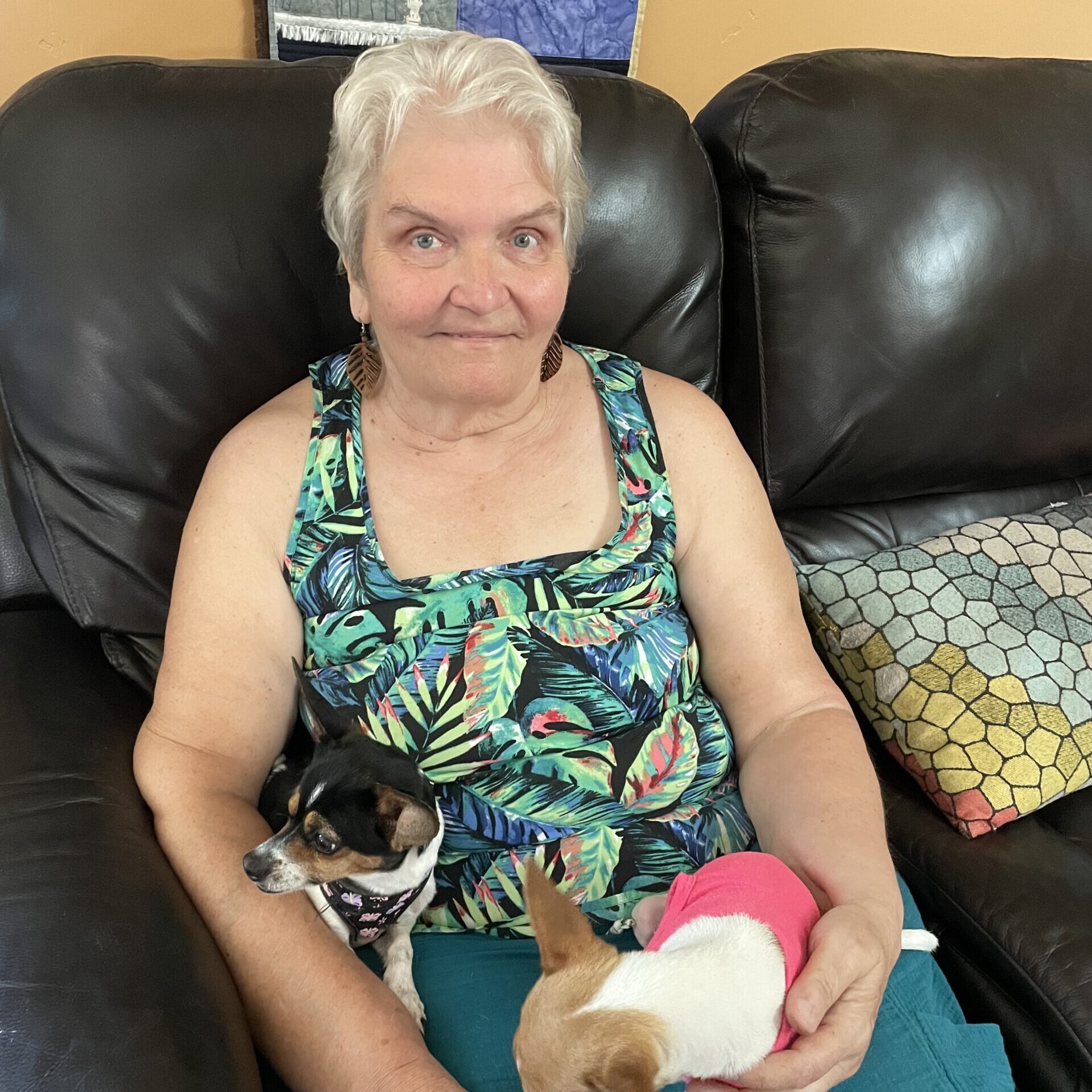 A white woman with short, white hair sits on a black couch with two small dogs in her lap. She wears a tank top with a leaf pattern and turquoise pants.