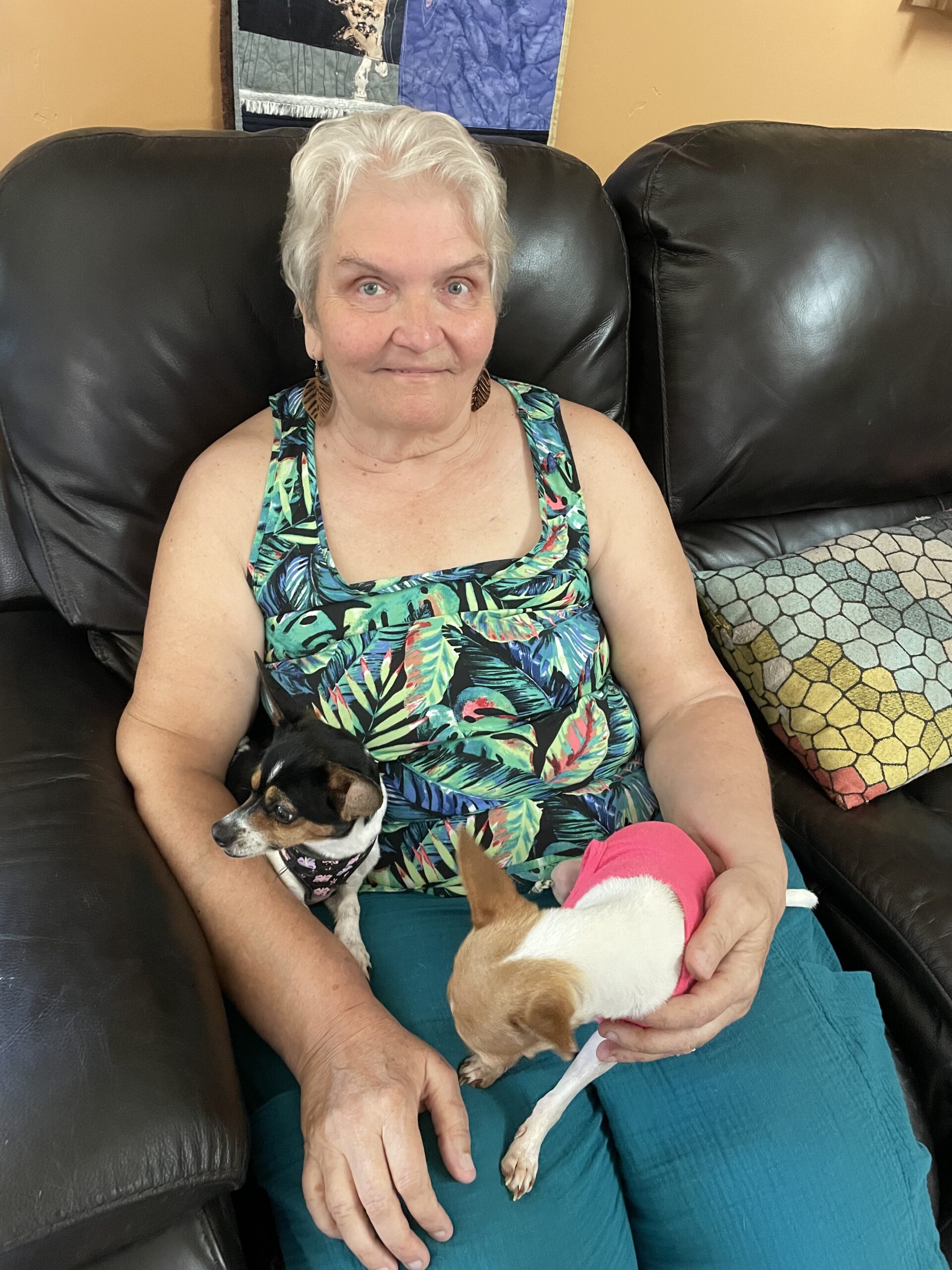A white woman with short, white hair sits on a black couch with two small dogs in her lap. She wears a tank top with a leaf pattern and turquoise pants.