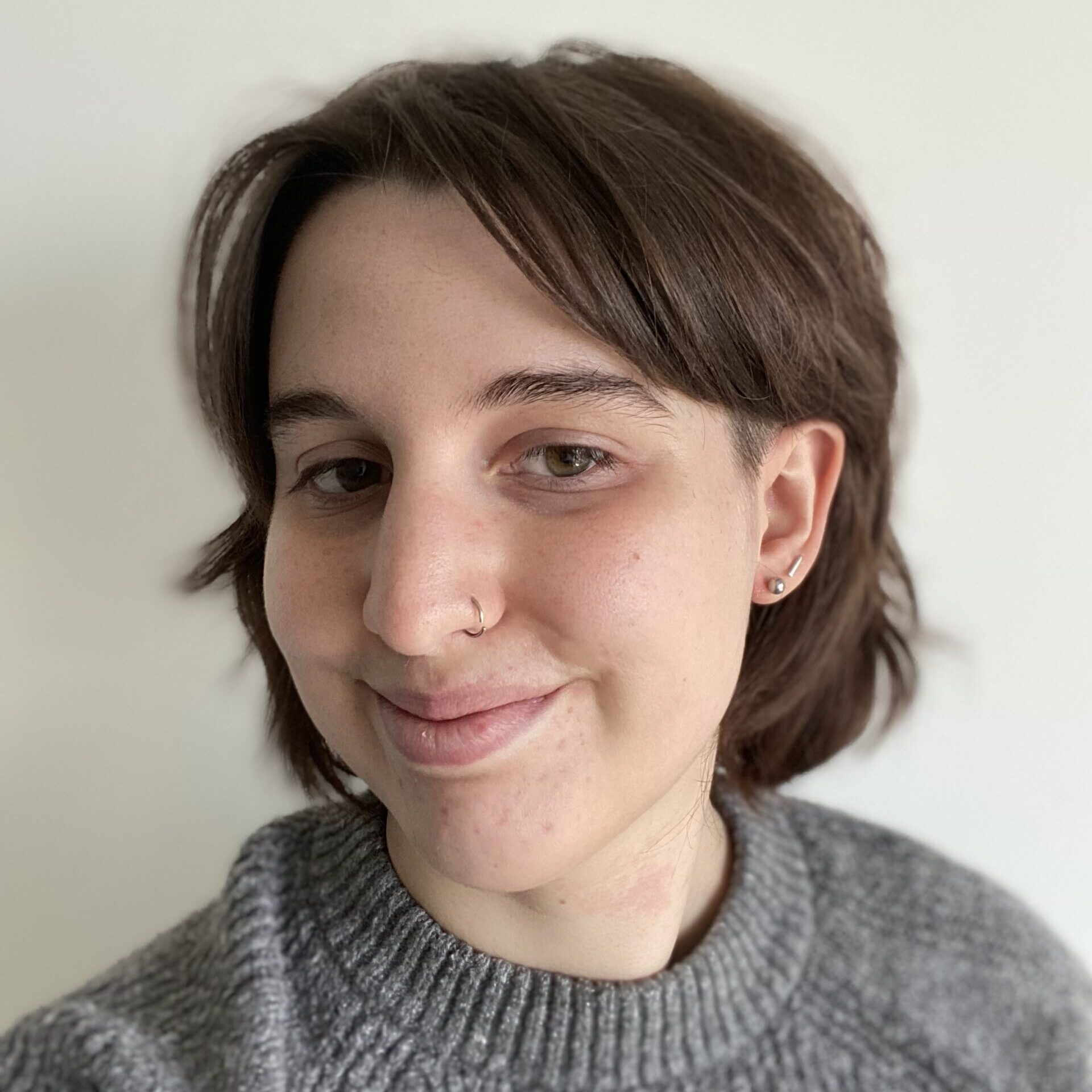 A white nonbinary person with short, brown hair wearing a gray sweater. They smile with their mouth closed and have a nose piercing.