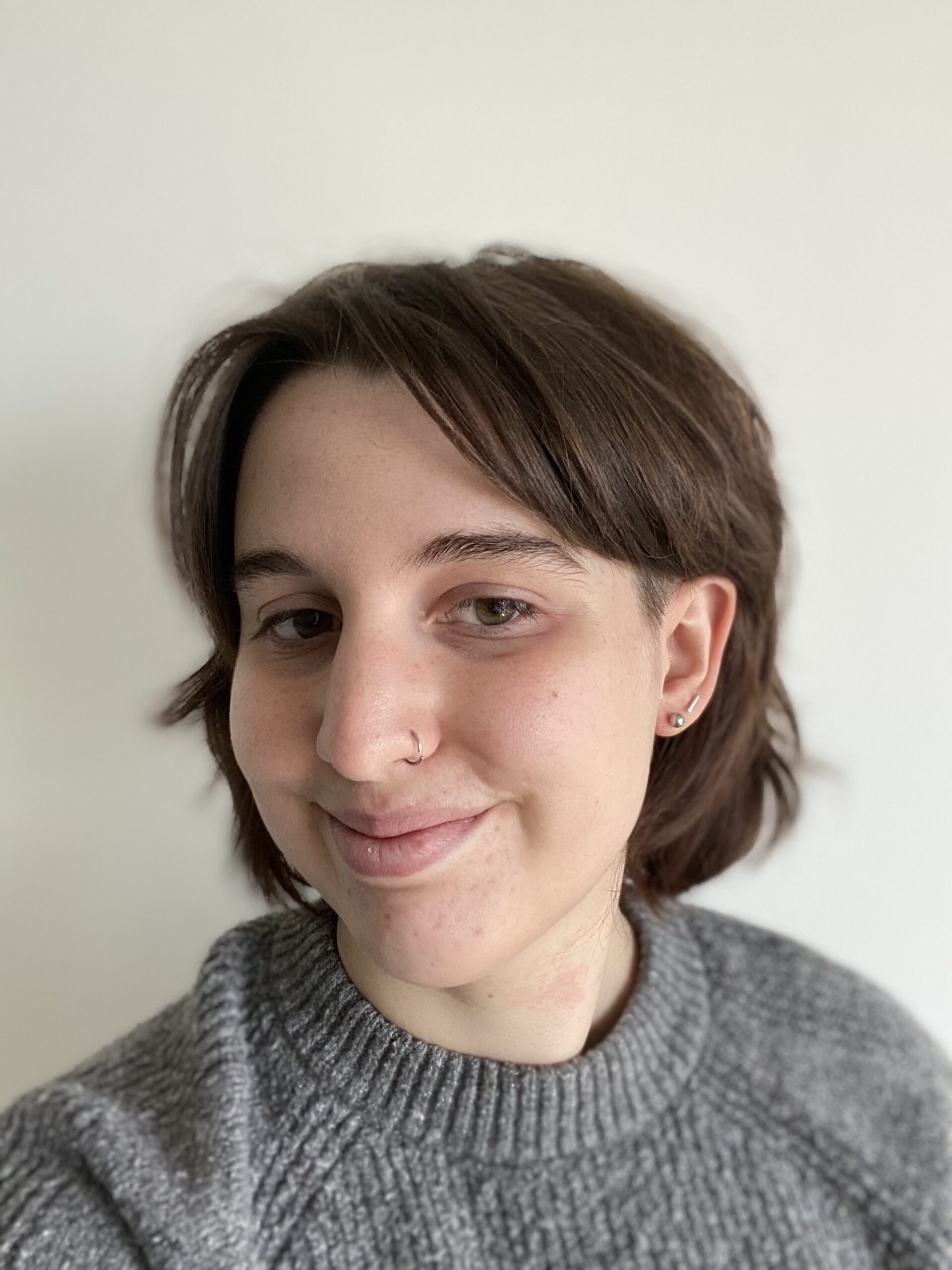 A white nonbinary person with short, brown hair wearing a gray sweater. They smile with their mouth closed and have a nose piercing.