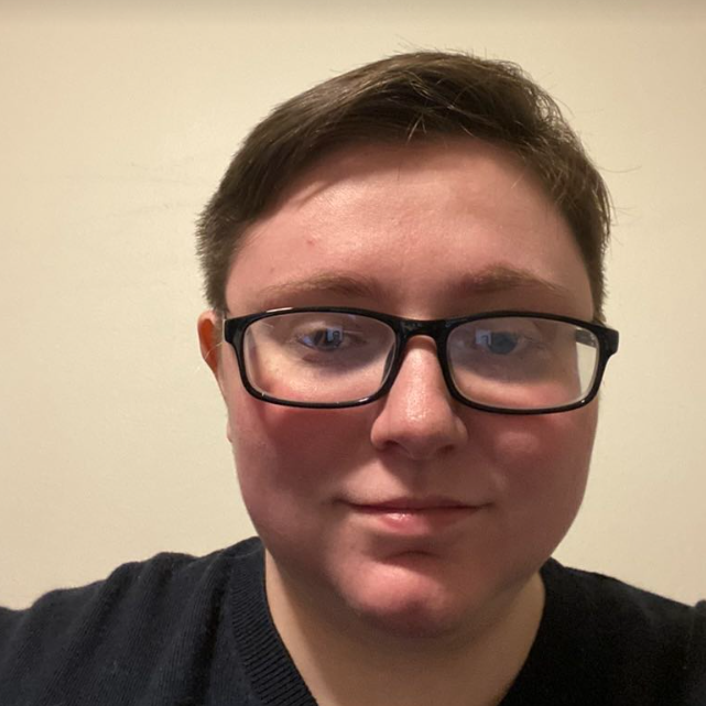 A white person with short, light brown hair and black, rectangular glasses smiles with their mouth closed. They wear a black T-shirt. The background is cream colored.
