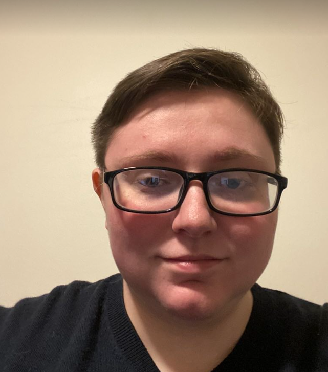 A white person with short, light brown hair and black, rectangular glasses smiles with their mouth closed. They wear a black T-shirt. The background is cream colored.