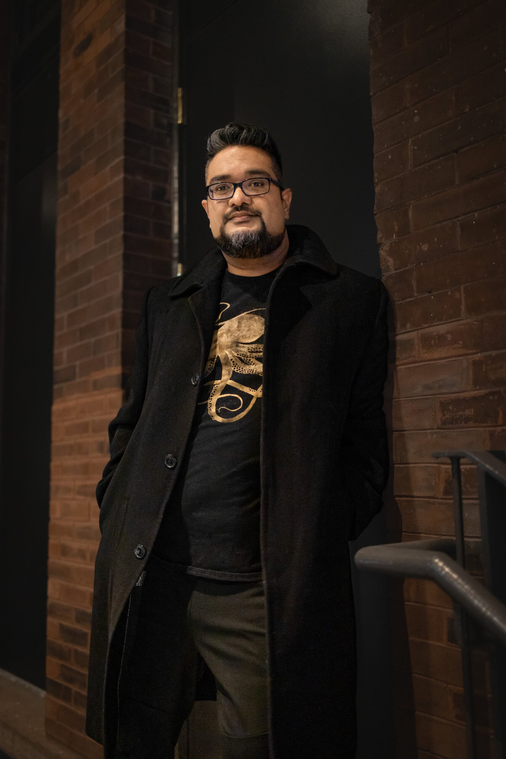 A man with brown skin, glasses, dark black hair, and a goatee. He stands against the railing in front of a brick building.