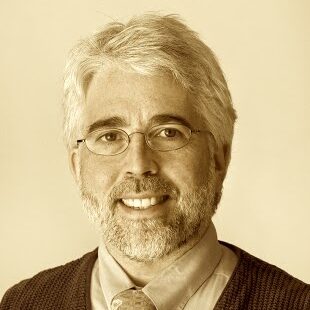 A sepia photo of a white man with white hair, a goatee, and wire-frame glasses. He wears a suit and tie and is smiling at the camera.