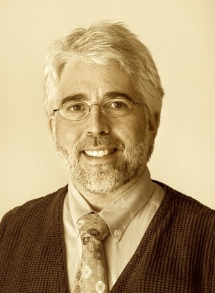A sepia photo of a white man with white hair, a goatee, and wire-frame glasses. He wears a suit and tie and is smiling at the camera.